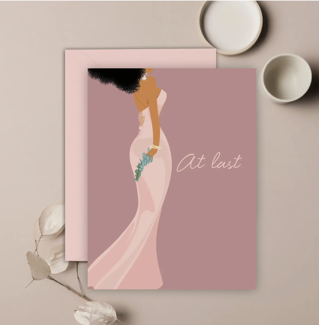 A mauve-colored greeting card depicting a stylized woman in a strapless wedding dress, with 'At last' written on it, accompanied by a matching envelope, presented on a neutral background.