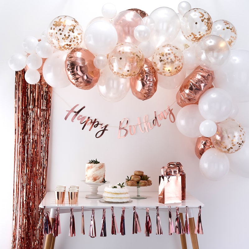 an arch of rose gold, cream, white, blush, and confetti filled balloons over a dessert table