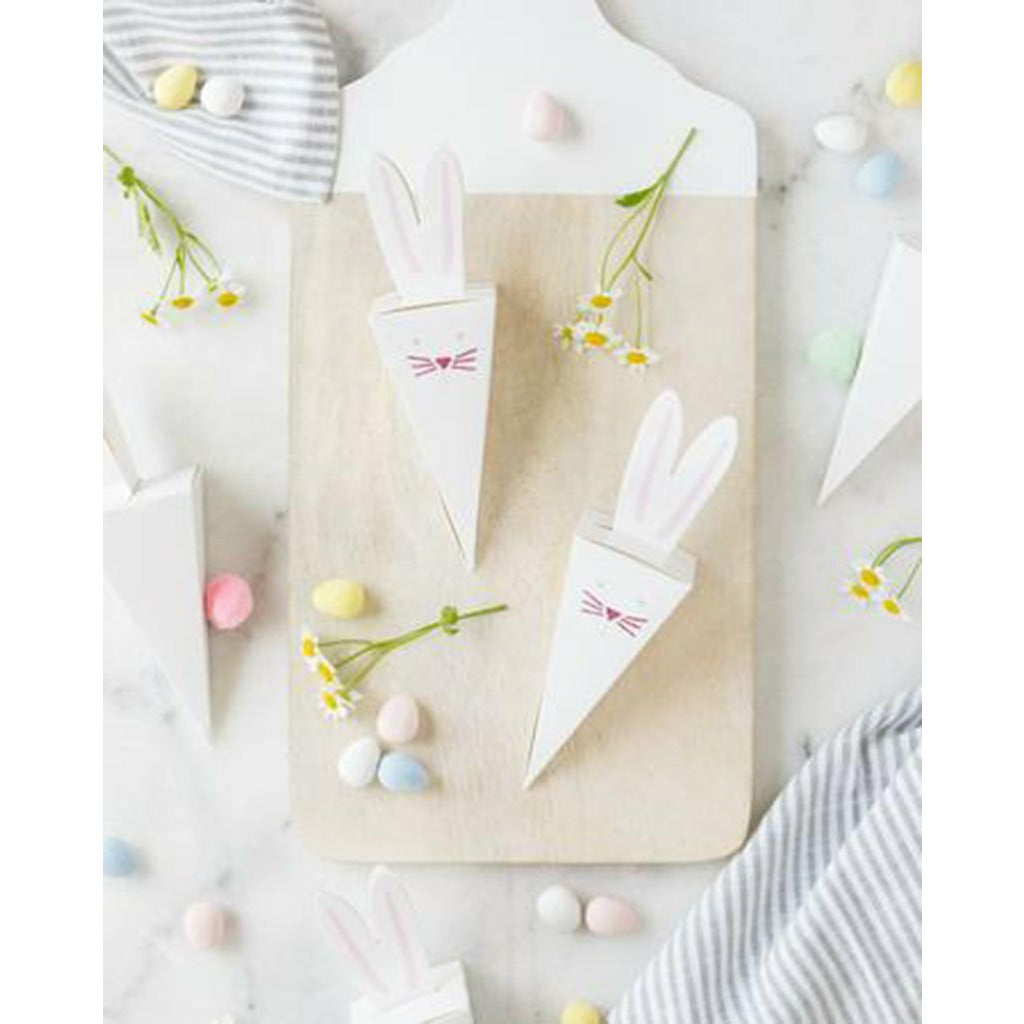 5 White triangle-shaped treat boxes with die cut bunny ears and pink printed bunny face surrounded jelly beans and candy eggs on a marble top