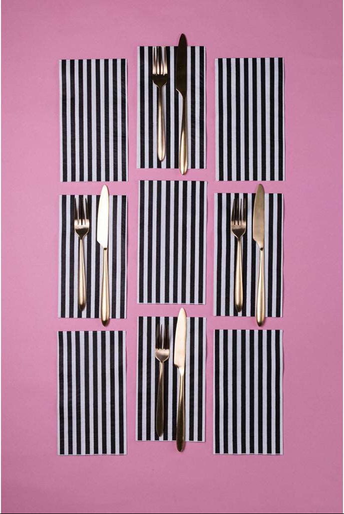 3 rows of Black &amp; White Striped Guest Napkins, a set of gold forks and knives on top of 4 of the napkins against a pink background