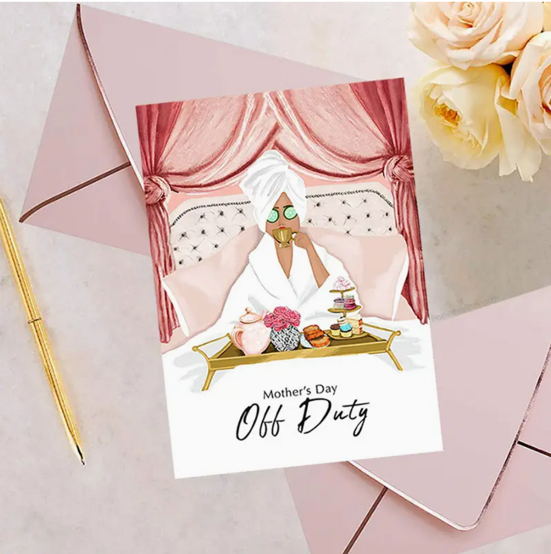Greeting card featuring an illustration of a woman in a spa robe and turban sitting on a pink upholestered bed, sipping tea from a gold cup, with a gold tray topped with pink roses and dessert platters. 2 pink envelopes with rose gold foil trim, gold pen, and cream and blush flowers