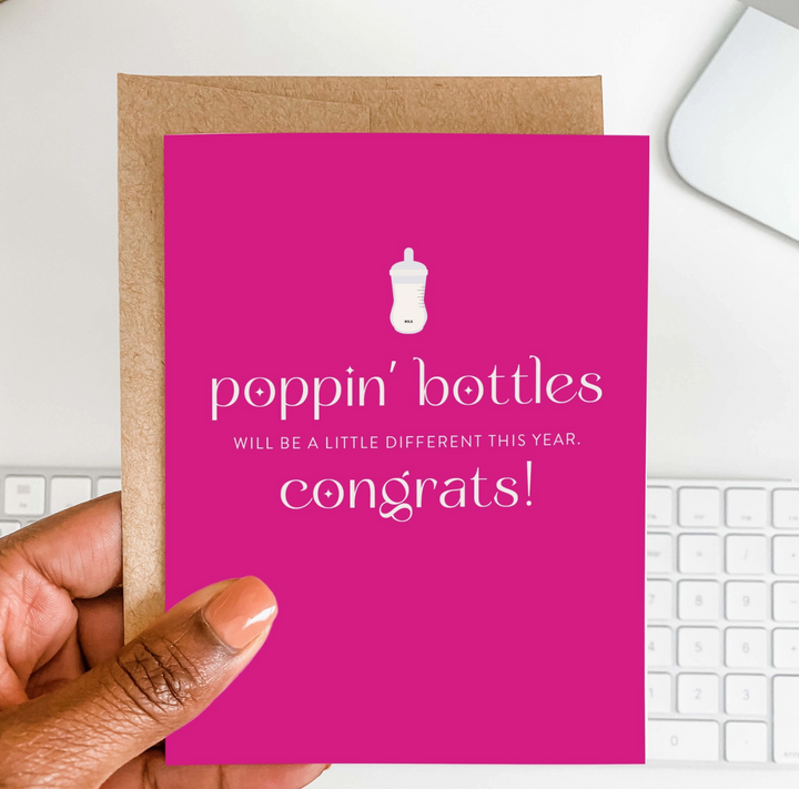 A hand holding a hot pink greeting card with white text that reads poppin bottles will be a little different this year. Congrats! and bottle icon, with brown envelope with a white keyboard in teh background
