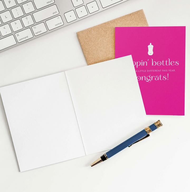 Hot pink greeting card with white text that reads poppin bottles will be a little different this year. Congrats! and bottle icon, with brown envelope, an open greeting card showing blank pages, a pen, and a partial view of a keyboard.