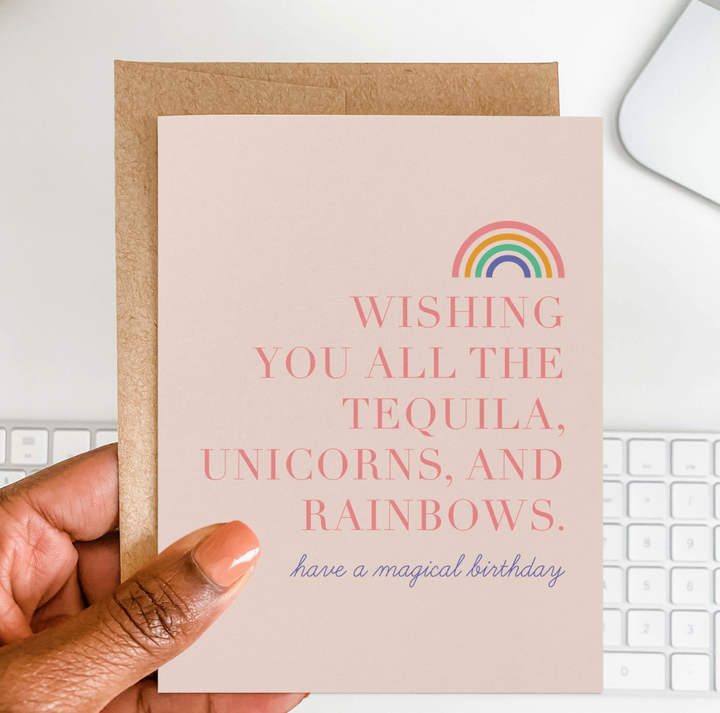 Hand holding a pink greeting card with a rainbow sketch which reads "wishing you all the tequila, unicorns, and rainbows. have a magical birthday" and a brown envelope with a white keyboard in the background