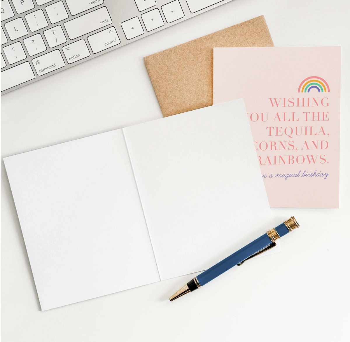 A pink greeting card with a rainbow sketch which reads "wishing you all the tequila, unicorns, and rainbows. have a magical birthday" and a brown envelope, an open greeting card showing blank pages, a pen, and a partial view of a keyboard.