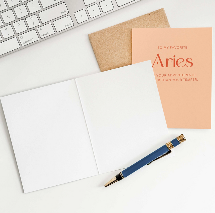 Peach greeting card with the text "to my favorite Aries, may your adventures be wilder than your temper" with a brown envelope, an open greeting card showing blank pages, a pen, and a partial view of a keyboard.
