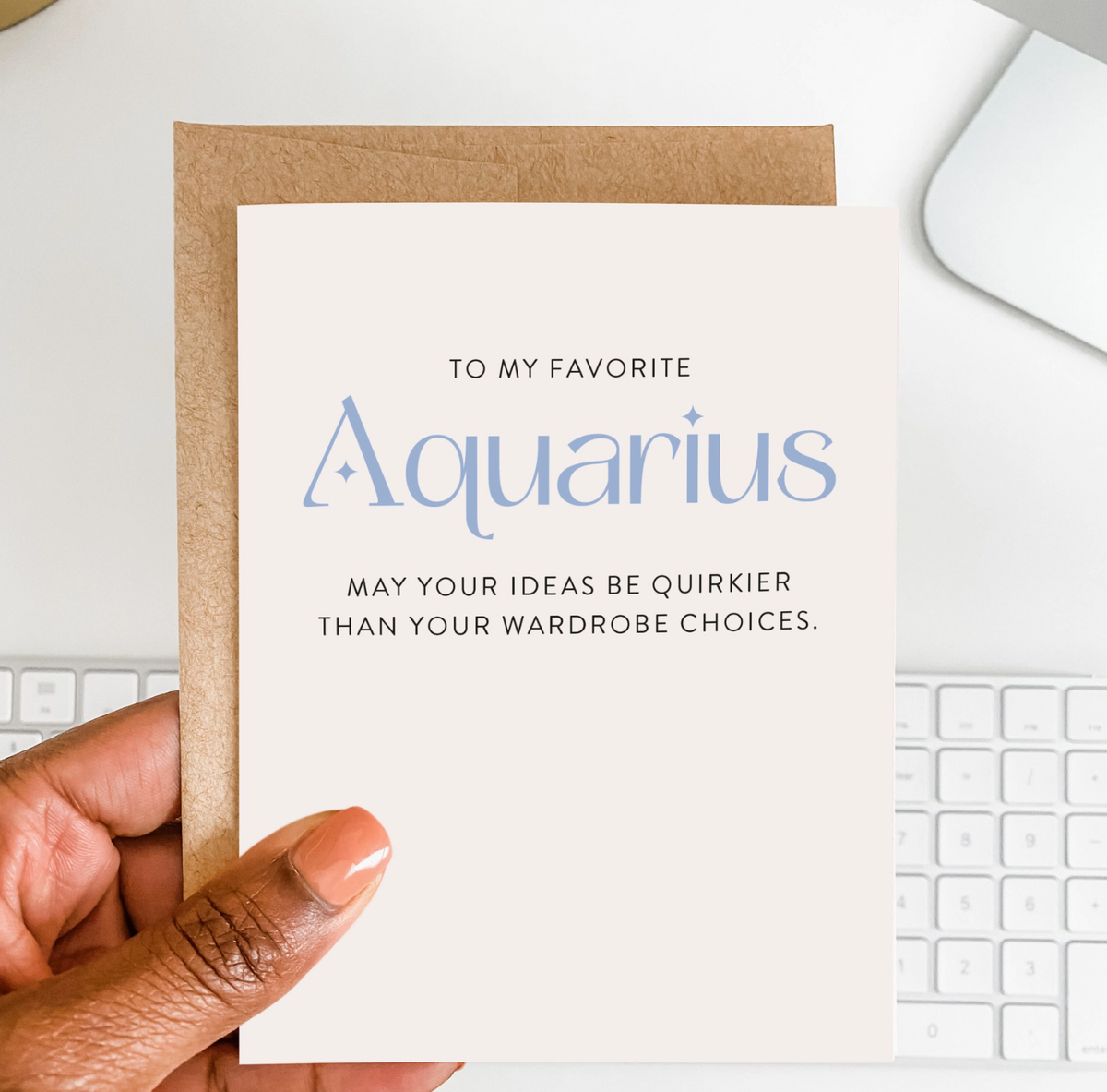 A card with text 'To my favorite Aquarius, may your ideas be quirkier than your wardrobe choices.' and a brown envelope held by a hand above a white keyboard