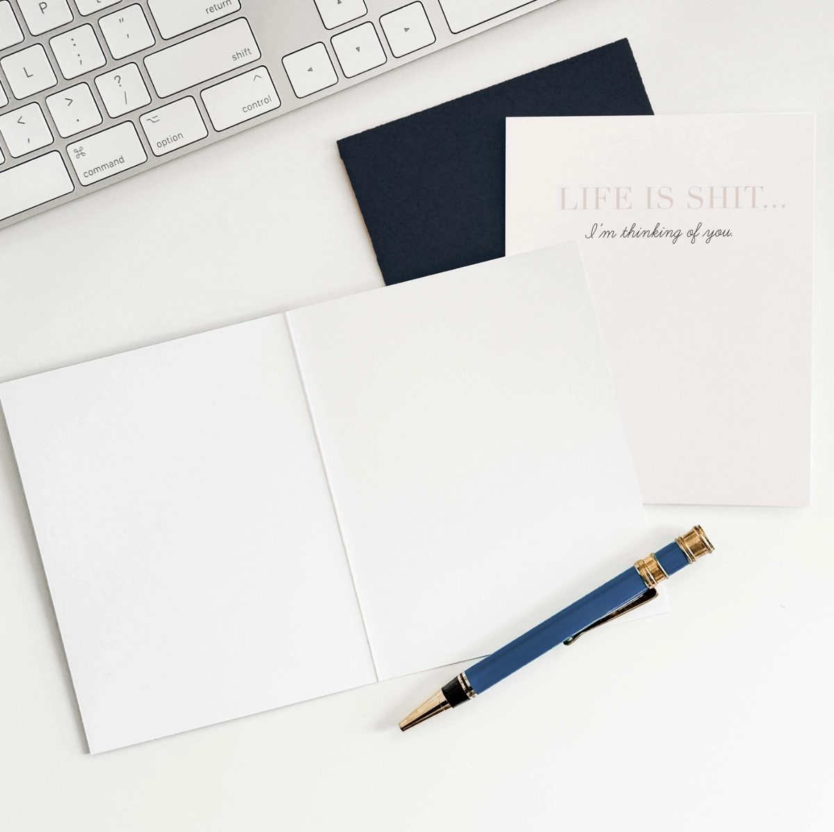 White greeting card with text 'Life is shit... I'm thinking of you' with a black envelope, an open greeting card showing blank pages, a pen, and a partial view of a keyboard.