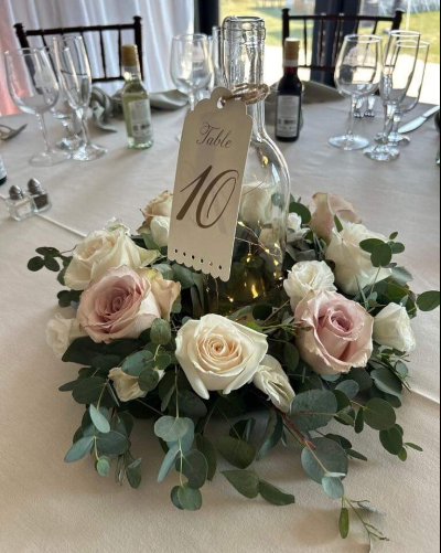 Floral centerpiece with a table number tag hanging from a clear glass wine bottle on a decorated table.