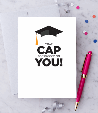White greeting card with a black graduation cap with gold tassel graphic and text that reads "That Cap Looks Good on You!" Paired with a metallic silver envelope