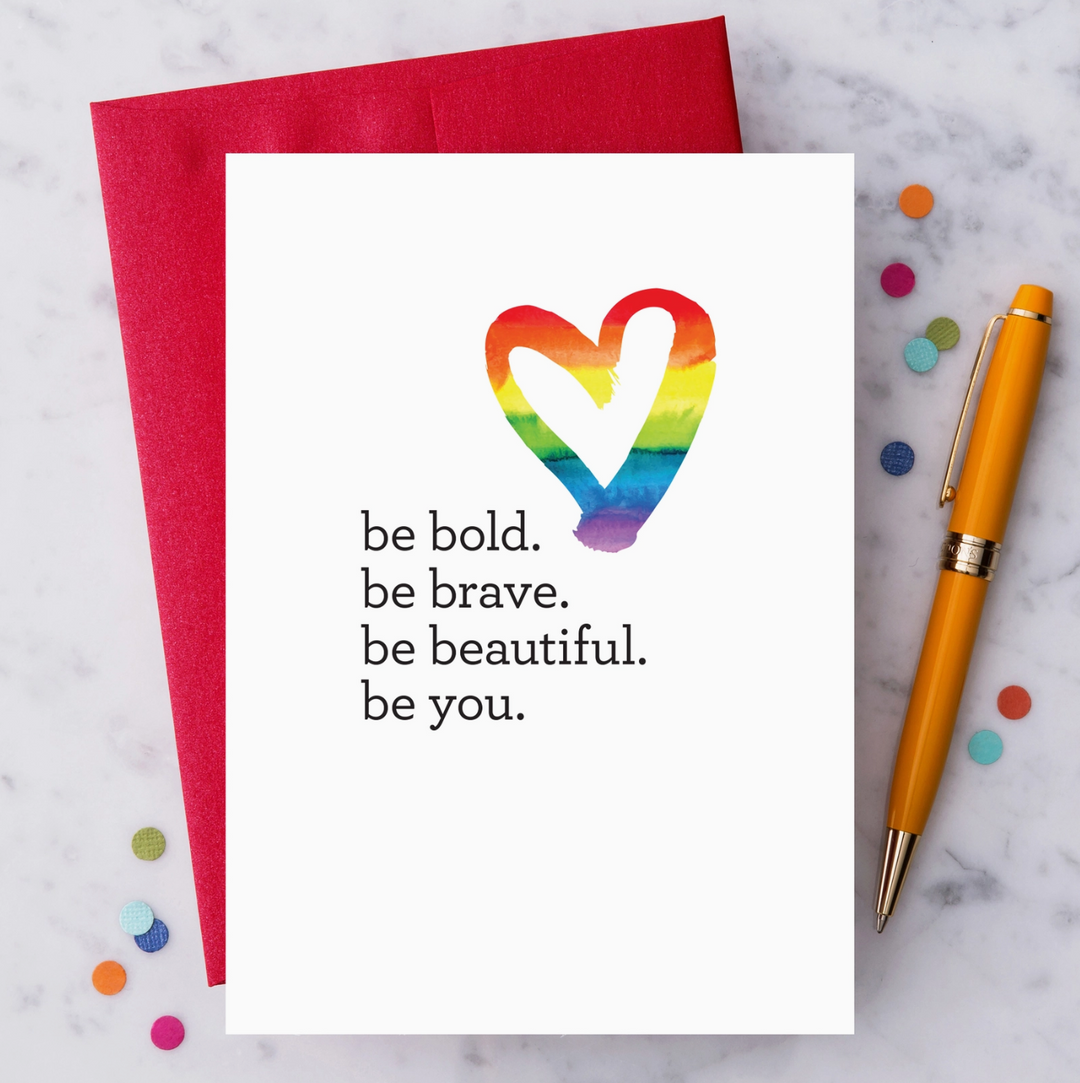 White greeting card with a rainbow striped heart graphic and text that reads "be bold. be brave. be beautiful. be you." on a marble surface with a pen and colorful paper confetti.