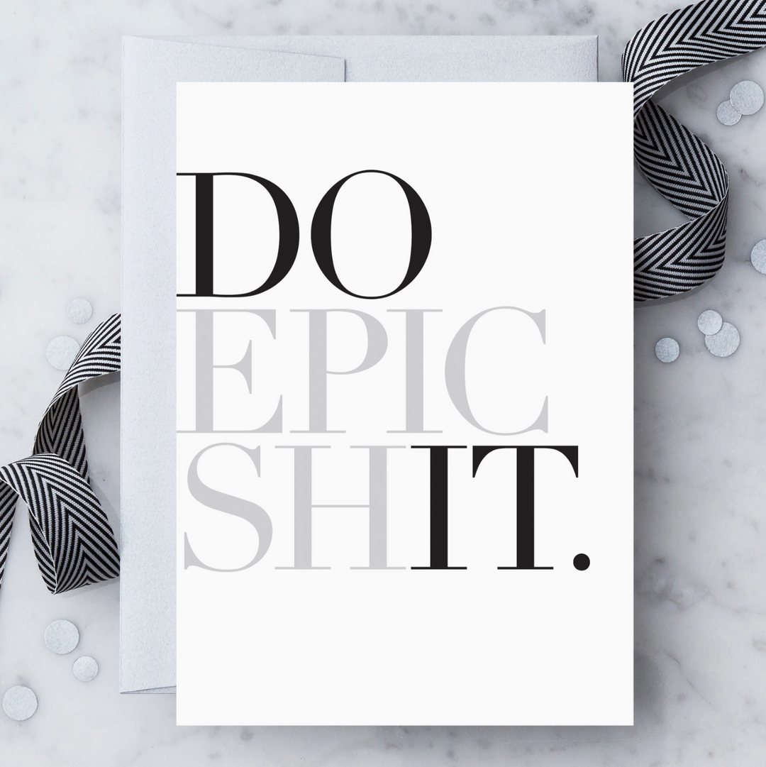 A white greeting card with black and metallic silver text that reads "DO EPIC SHIT." Paired with a silver envelope on a light marble surface with a black-and-white ribbon and silver confetti.