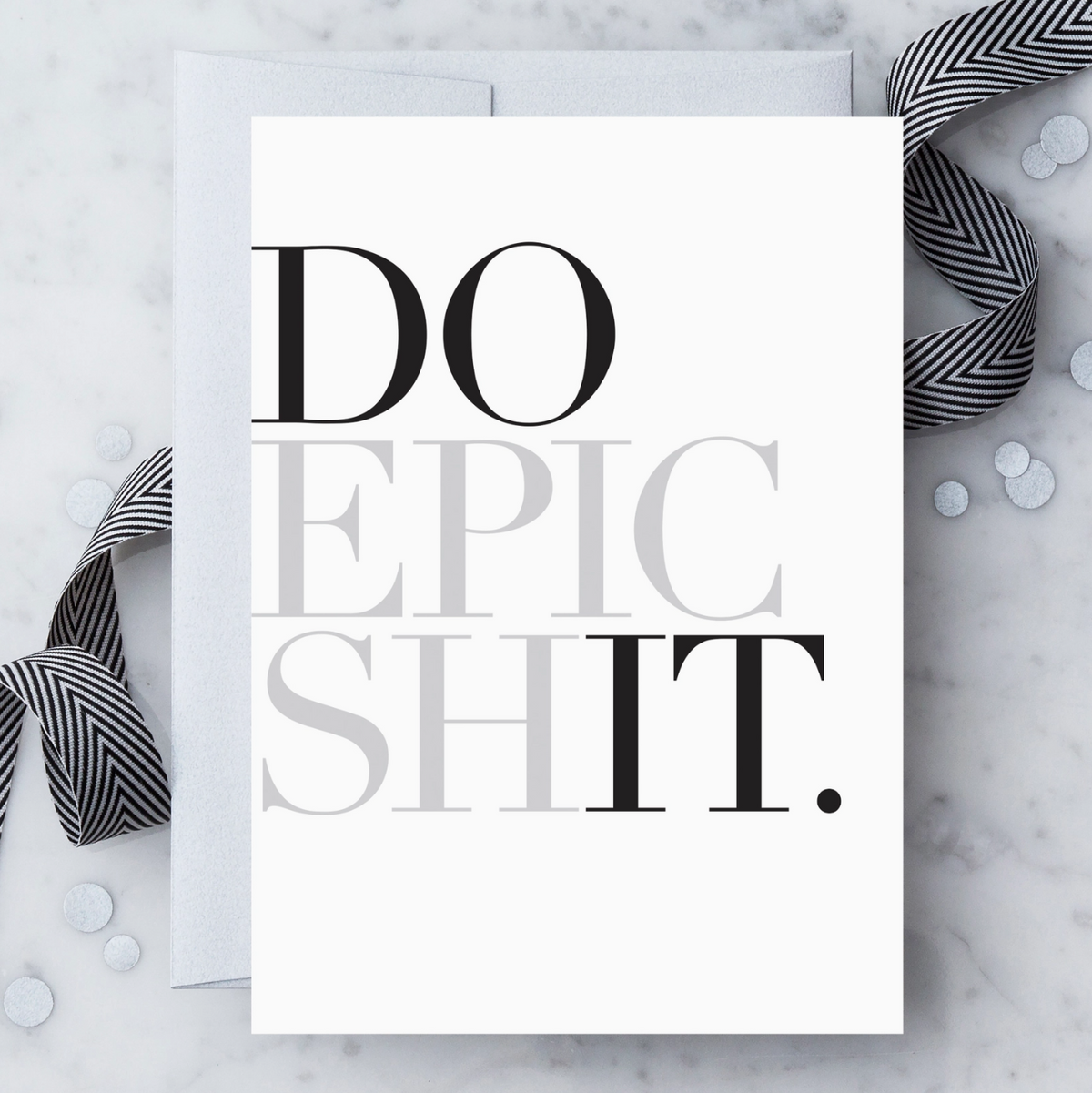 A white greeting card with black and metallic silver text that reads "DO EPIC SHIT." Paired with a silver envelope on a light marble surface with a black-and-white ribbon and silver confetti.