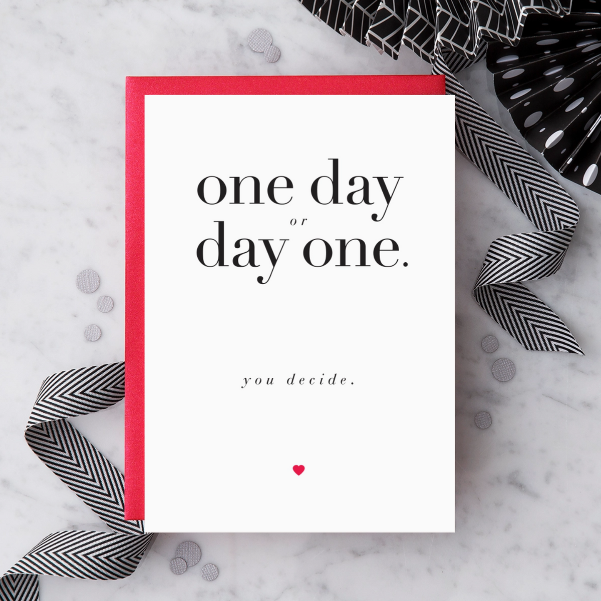 White greeting card with black text that reads "one day or day one. you decide." and a red heart below text. Paired with a red envelope on a marble background with black and white decorative objects