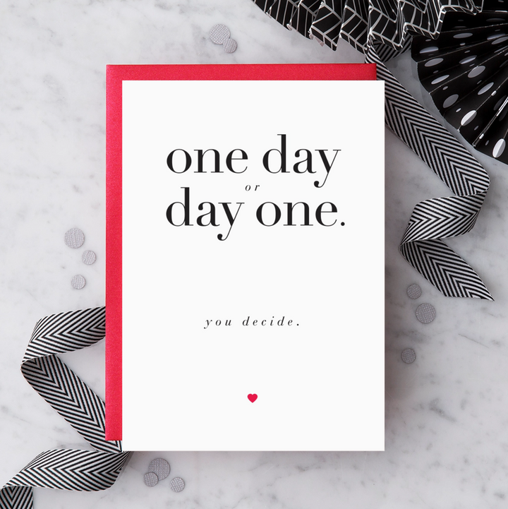 White greeting card with black text that reads "one day or day one. you decide." and a red heart below text. Paired with a red envelope on a marble background with black and white decorative objects