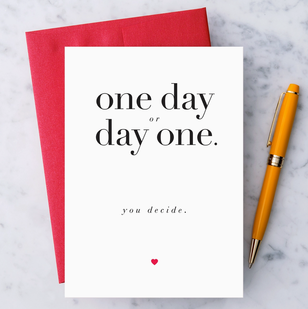 White greeting card with black text that reads "one day or day one. you decide." and a red heart below text. Paired with a red envelope on a marble background with a orange pen.