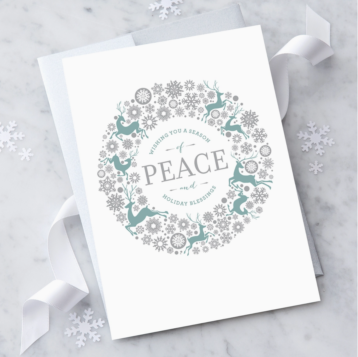 White greeting card, with an illustrated wreath made of a variety of silver snowflakes and green reindeers surround silver text that reads "Wishing you a season of peace and holiday blessings". Paired with a silver envelope on a marble background with white ribbon and snowflake shaped confetti. 