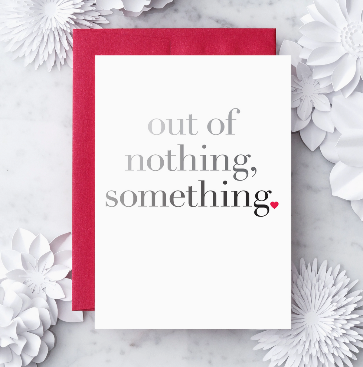 White greeting card with a grey to black gradient text that reads "out of nothing, something" and a red heart shaped graphic positioned as a period. Paired with a red envelope on a white marble background surrounded by white decorative objects.