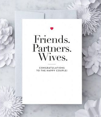 White greeting card with black text that reads "Friends. Partners. Wives. Congratulations to the happy couple!" with a red heart graphic. Paired with a metallic silver envelope on a marble background