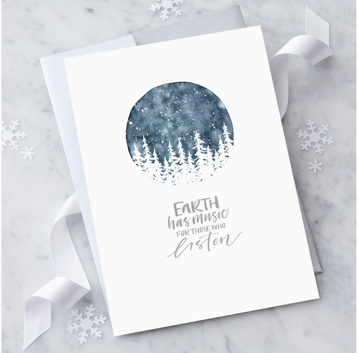 White greeting card with silver text that reads "Earth has music for those who listen" with an illustration of a midnight snow filled sky and white christmas trees. Paired with a silver envelope on a marble background with white ribbon and snowflake shaped confetti