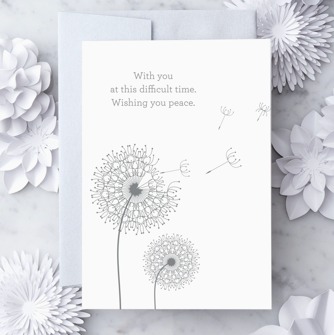 White greeting card with metallic silver text that reads "With you at this difficult time. Wishing you peace." with a metallic silver illustration of 2 clovers. Paired with a silver envelope on a marble background with decorative objects