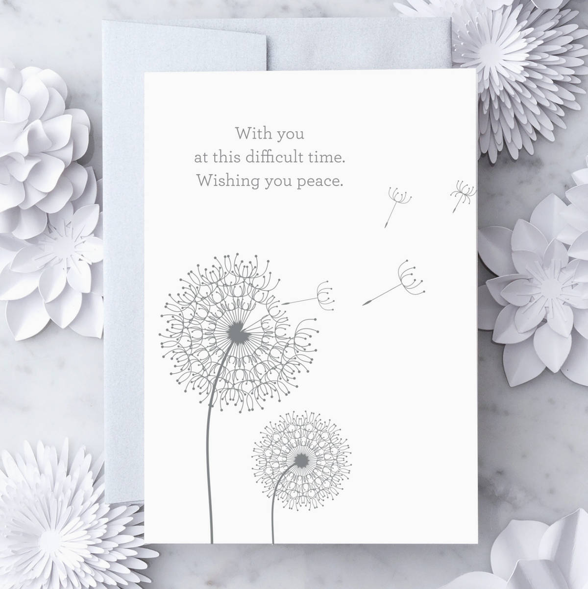 White greeting card with metallic silver text that reads "With you at this difficult time. Wishing you peace." with a metallic silver illustration of 2 clovers. Paired with a silver envelope on a marble background with decorative objects