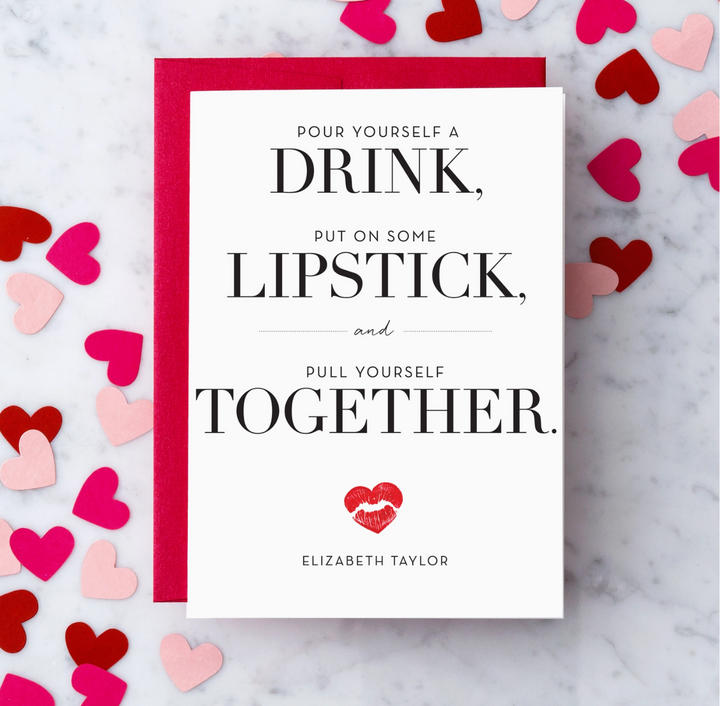 White greeting card with black text that reads "Pour yourself a drink, put on some lipstick, and pull yourself together. Elizabeth Taylor" with a heart shaped kiss graphic in red lipstick. Paired with a red envelope on a marble background surrounded by red, blush pink and hot pink heart shaped confetti