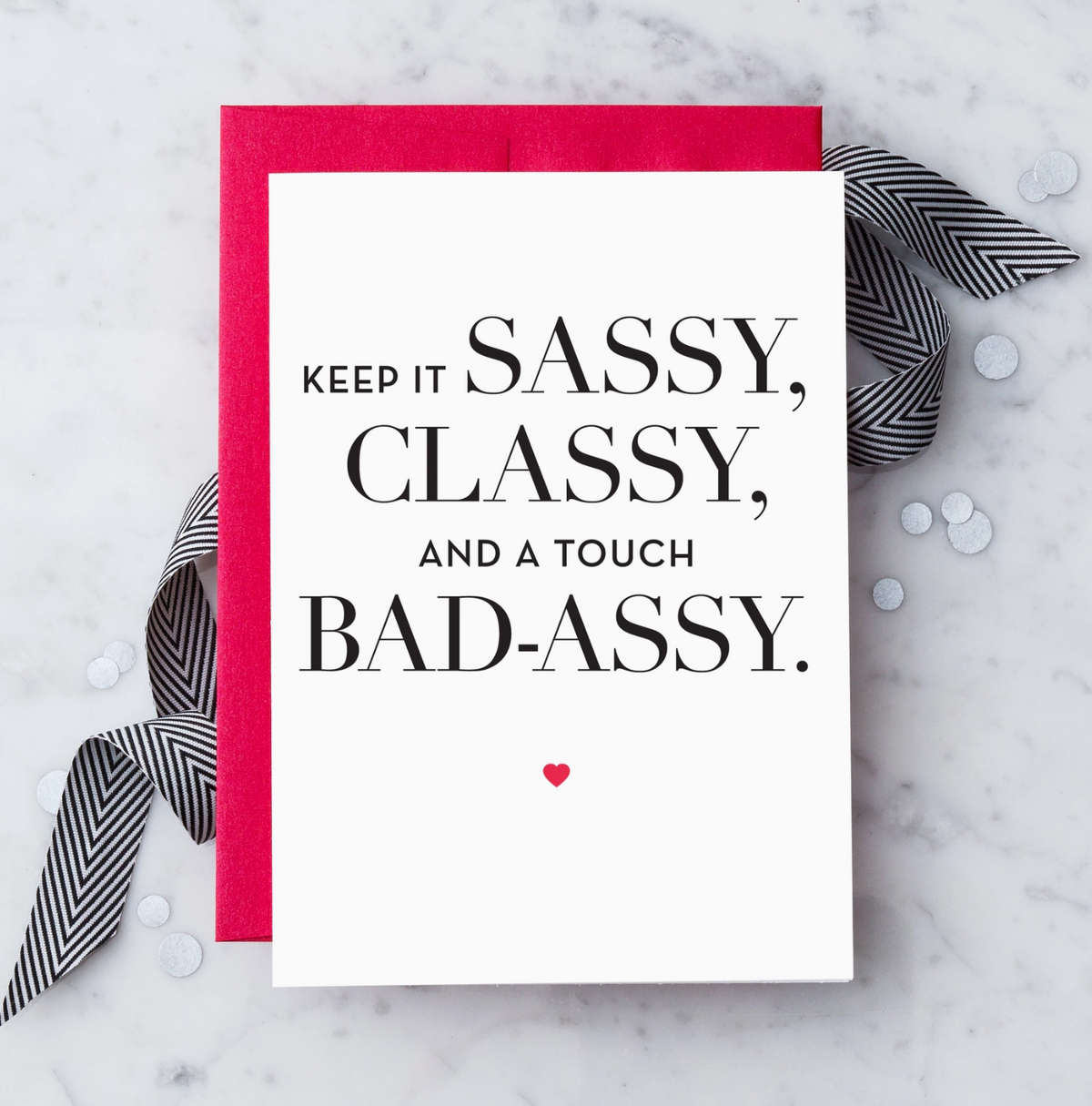 A white greeting card with black text reading "KEEP IT SASSY, CLASSY, AND A TOUCH BAD-ASSY." and a small red heart. Paired with a red envelope on a marble background with a black and white ribbon and white confetti