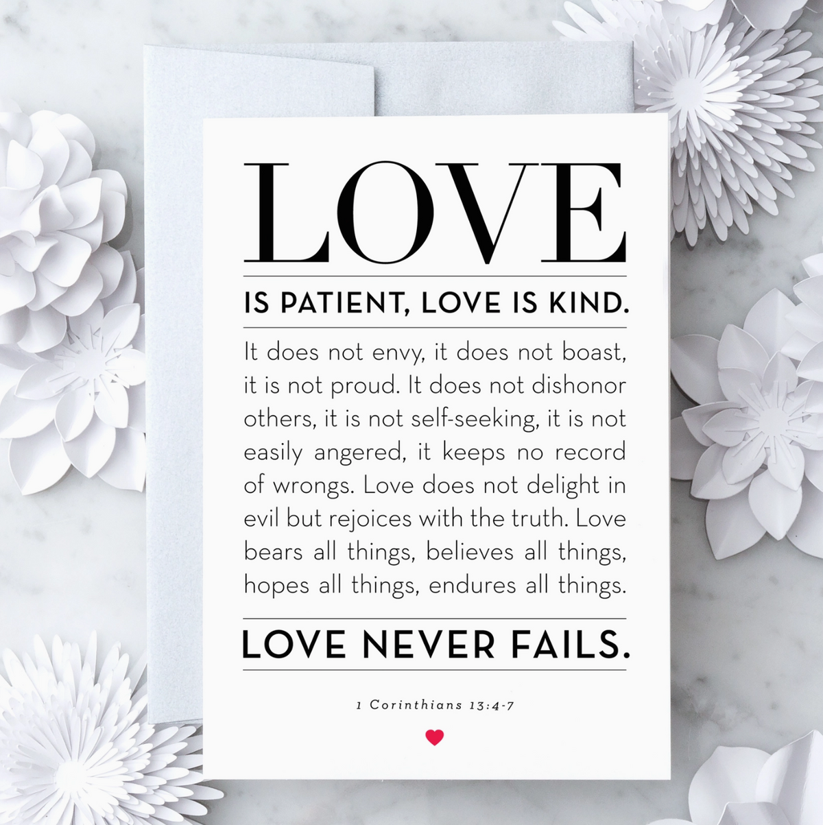 White greeting card with black text that reads "Love is patient, love is kind. it does not envy, it does not boast, it is not proud. it does not dishonor others, it is not self-seeking, it is not easily angered, it keeps no record of wrongs. Love does not delight in evil but rejoices with the truth. Love bears all things, believes all things, hopes all things, endures all things. Love never fails. 1 Corinthians 13:4-7" with a red heart graphic. Paired with a metallic silver envelope on a marble background