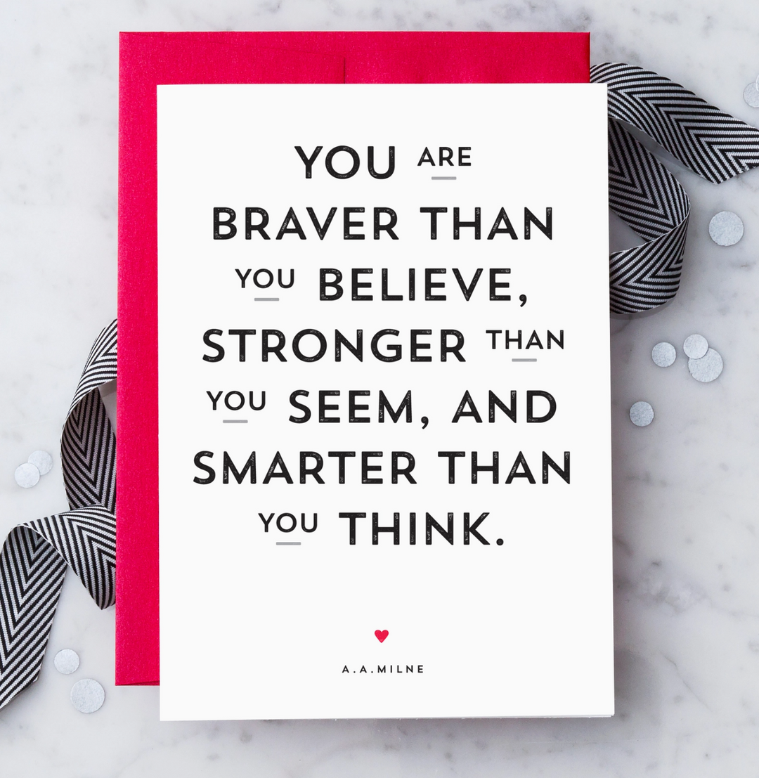 A white typographic greeting card with a quote in black text reading, "YOU ARE BRAVER THAN YOU BELIEVE, STRONGER THAN YOU SEEM, AND SMARTER THAN YOU THINK." with a small red heart and the name A.A. Milne. The card is paired with a red envelope on a marble background with black and white ribbon and confetti