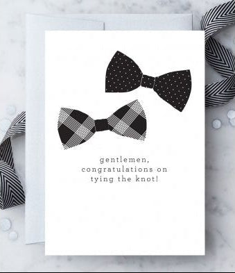 White greeting card with 2 black-and-white bowtie illustrations and text that reads "Gentlemen, Congratulations on tying the knot!" Paired with a metallic silver envelope on a marble background, with white confetti and a black-and-white ribbon accent.