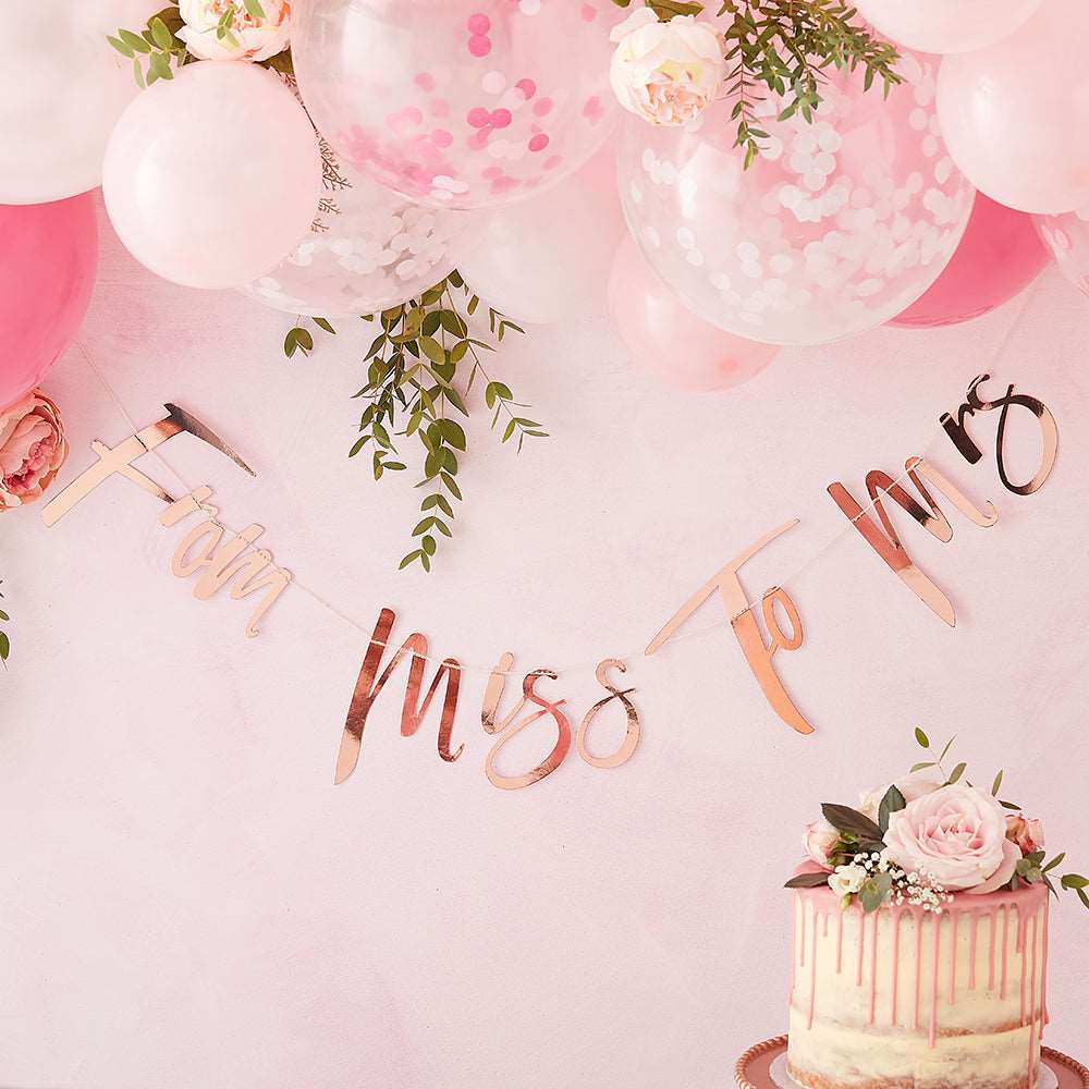 From Miss. to Mrs. Letter Banner against a pink wall, white and pink ballons and foliage hanging from ceiling and a flower topped wedding cake
