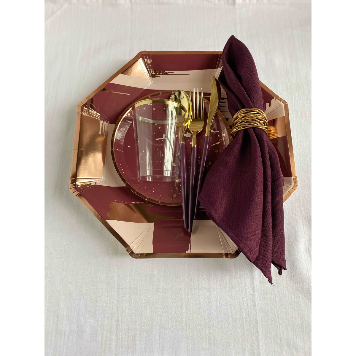 top view of a table setting, linen colored tablecloth, mauve, blush pink, and rose gold brushstroke print patterned hexagon-shaped charger plate, burgundy dessert plate with rose gold rim and rose gold and white splatter print pattern, purple handled gold utensils, purple cloth napkin with gold napkin ring, and a clear plastic cup with gold rim
