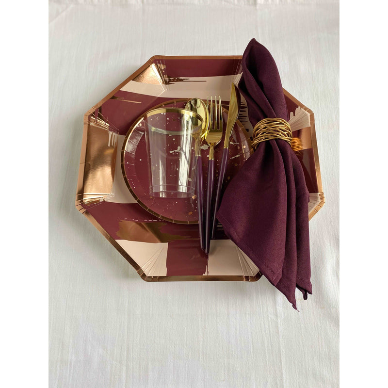 top view of a table setting, linen colored tablecloth, mauve, blush pink, and rose gold brushstroke print patterned hexagon shaped charger plate, burgundy dessert plate with rose gold rim and rose gold and white splatter print pattern, purple handled gold utensils, purple cloth napkin with gold napkin ring, and a clear plastic cup with gold rim