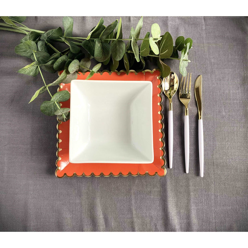 top view of a table setting grey tablecloth with spiral eucalyptus plant, gold rim, scalloped edge orange dinner plate topped with a square white dinner bowl, and white handled with gold utensils on the side