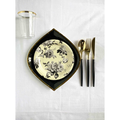 Top view of a white linen tablecloth, black gold rimmed dinner plate with an ivory color with black floral print dessert plate and a black handled and gold cutlery set and a clear cup with gold rim.
