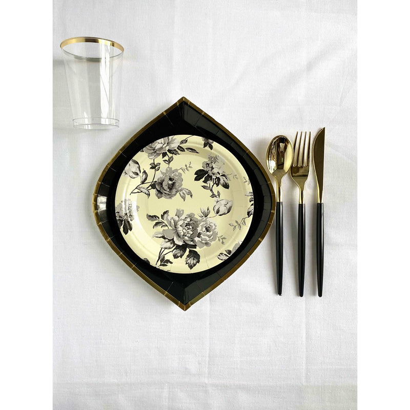 Top view of a white linen tablecloth, black gold rimmed dinner plate with an ivory color with black floral print dessert plate and a black handled and gold cutlery set and a clear cup with gold rim.