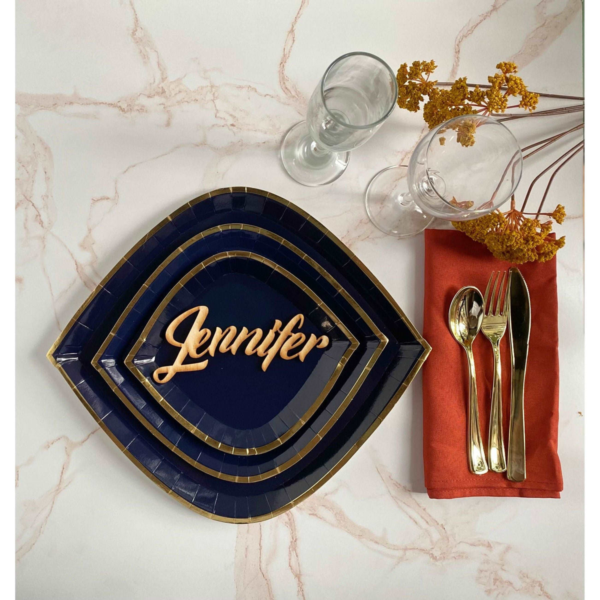 marble tabletop with navy, gold rimmed plate set (charger, dinner, and dessert), 2 clear wine glasses, an orange cloth napkin and a gold utensil set