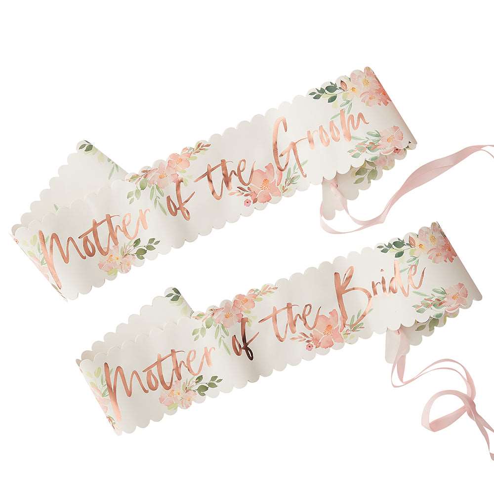 Two paper sashes with floral patterns and 'Mother of the Bride and Mother of the Groom' printed in rose gold foil, tied with pink ribbons.