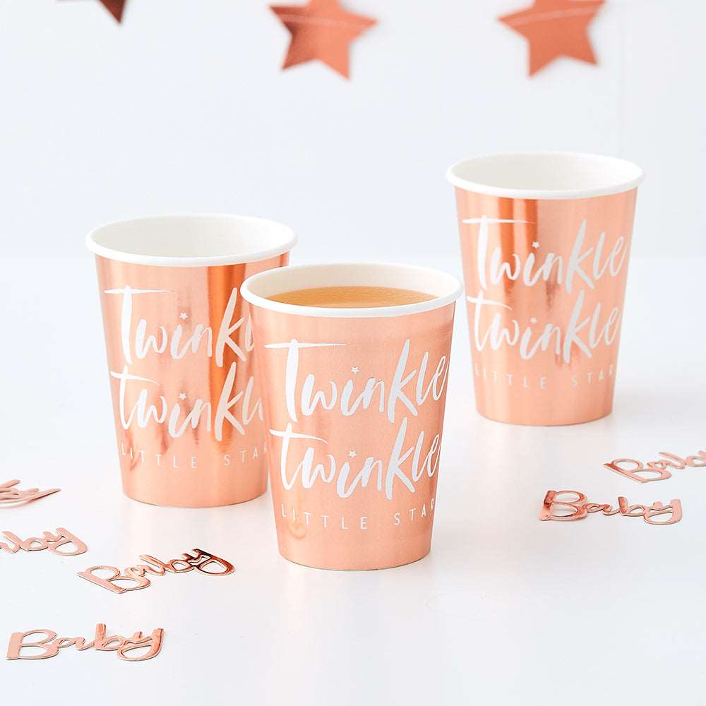 white tabletop with 3 rose gold foil paper cups with white script font that reads Twinkle Twinkle, and a regular font that reads little star, rose gold foil confetti words scattered that read baby, rose gold die-cut star banner in the background