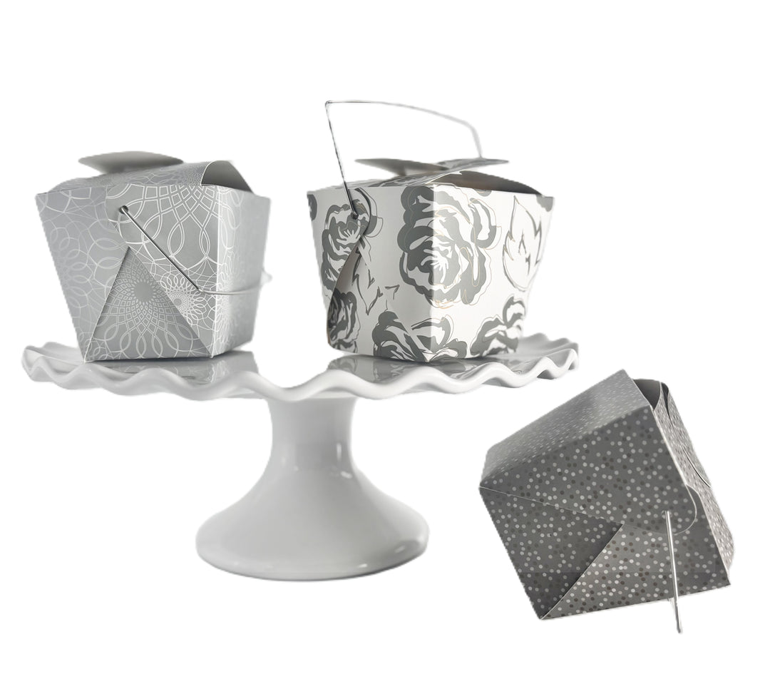 3 decorative take-out style boxes one with an allover silver and gold floral pattern, one silver box with an allover white scroll pattern on a white cake stand and one charcoal gray box with a tonal grey and gold polka dot pattern positioned next to the cake stand.