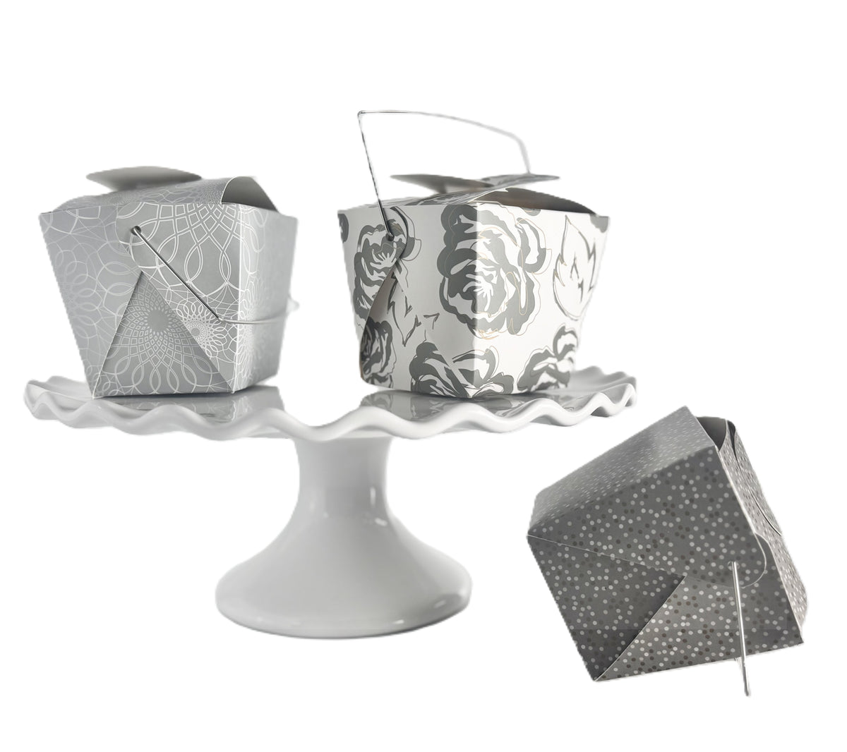 3 decorative take-out style boxes one with an allover silver and gold floral pattern, one silver box with an allover white scroll pattern on a white cake stand and one charcoal gray box with a tonal grey and gold polka dot pattern positioned next to the cake stand.