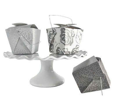 3 decorative take-out style boxes one with an allover silver and gold floral pattern, one silver box with an allover white scroll pattern on a white cake stand and one charcoal gray box with a tonal grey and gold polka dot pattern positioned next to the cake stand.