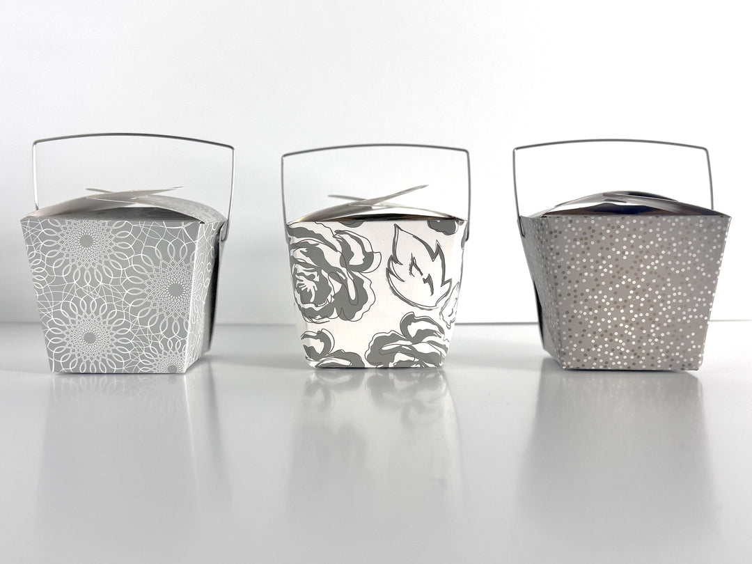 3 decorative take-out style boxes next to each other. One with an allover silver and gold floral pattern, one silver box with an allover white scroll pattern, and one charcoal gray box with a tonal grey and gold polka dot pattern against a white background.