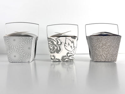 3 decorative take-out style boxes next to each other. One with an allover silver and gold floral pattern, one silver box with an allover white scroll pattern, and one charcoal gray box with a tonal grey and gold polka dot pattern against a white background.