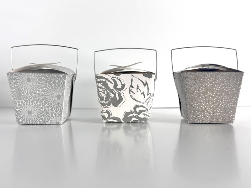 3 decorative take-out style boxes next to each other. One with an allover silver and gold floral pattern, one silver box with an allover white scroll pattern, and one charcoal gray box with a tonal grey and gold polka dot pattern against a white background.