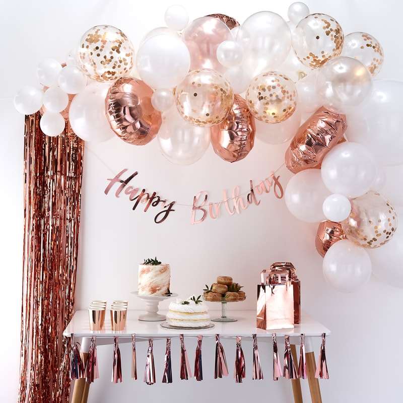A rose gold, matte white, pearl white, and clear with rose gold confetti-filled balloon arch, rose gold streamer-style curtain, a rose gold happy birthday letter banner, all against a white wall above a dessert table.