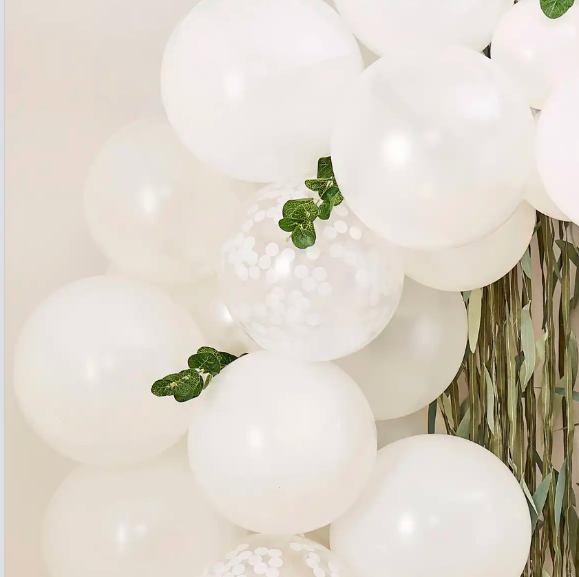 A combination of solid white and clear white confetti-filled balloons with green eucalyptus sprigs against a grass wall