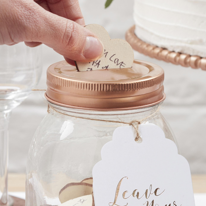 Hand placing a wooden heart with special messgae written on it into a glass jar with a copper lid, surrounded by wine glasses and cake on cake stand