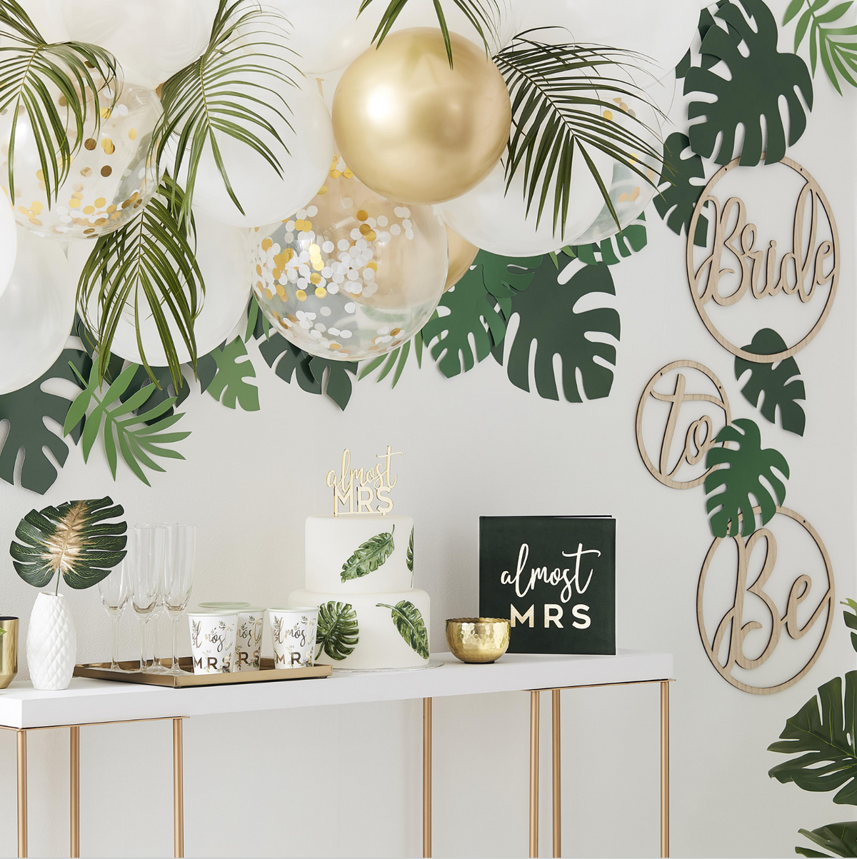 Tropical-themed bridal shower setup with matte white, pearl white, gold, and clear gold confetti-filled balloon arch with green foliage accents. Faux monstera leaves and circular wooden bride to be signs on a white wall above a gold and white beverage table.